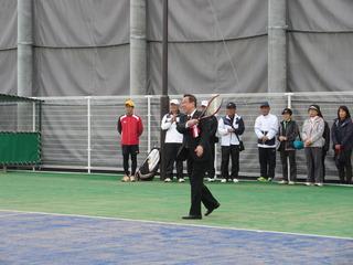テニスコートで胸に紅白の胸章を付けた市長がテニスラケットを振っている写真