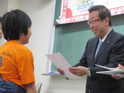 「子ども大学ふじみの」の修了式で、オレンジ色のティーシャツを着た男の子が市長から修了証書を読まれている写真