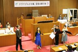 議長席の前で、フルート演奏者の古田 土明歌さんや他の演奏者の方々が楽器を持って写している写真