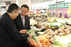 店に沢山のじゃがいもやニンジン白菜などの野菜が並べられており、キュウリを手に持って、市長と男性が話をしている写真