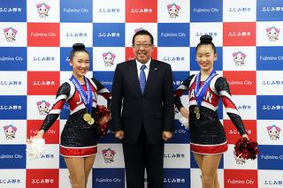 首からメダルを2個かけた新保 心海さんと村岡 虹音さんがチアリーディングのユニフォームを着て、ポーズを決めて市長と笑顔で写している写真