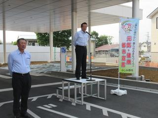 駐車場に設置された朝礼台に市長が立ってスタンドマイクの前で話をしている写真