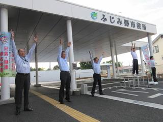 朝礼台に男性が立って見本の体操をしており、その横で市長と職員の方々が両手を耳に付けてまっすく挙げて体操をしている写真