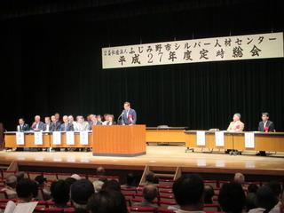 シルバー人材センター定時総会の会場の壇上で市長が話をしており、舞台上には長机が置かれ関係者の方々が座っている