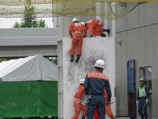 オレンジ色の服を着た消防隊員が白い高い壁を2人の隊員が足場を作って、別の2人の隊員が壁をよじ登っている写真