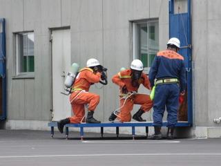 消防隊員が空気呼吸器のマスクを着けてボンベを背負っており、腰にロープを巻いて台の上で訓練をしている写真