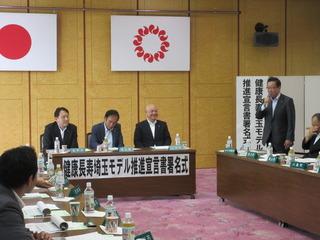 健康長寿埼玉モデル推進宣言署名式の会場にて市長が席から立ち上がってマイクを持って話をしており、関係者の方々は席に着いて話を聞いている写真