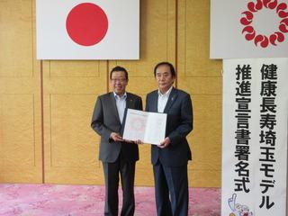 上田 清司埼玉県知事と市長が証書を手に持って「健康長寿埼玉モデル推進宣言署名式典式」と書かれている看板の横で写っている写真