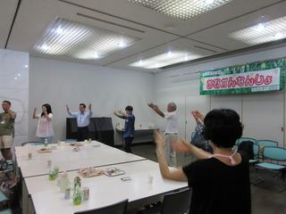参加者が机の周りで円になり、みんなで踊りを踊っている写真