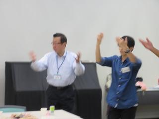 青いシャツをきた関係者の人に踊りを教わりながら手を挙げ踊っている市長の写真