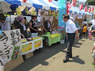 祭りの会場で奄美皇帝マンゴースムージーと書かれた出店の前を歩いている市長の写真