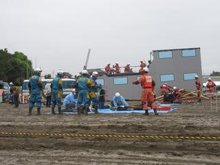 ブルーシートの上でマネキンを使って救助訓練をしている奥に倒壊した建物が写っている写真