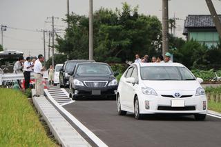 道路の両脇に関係者が並び拍手をしているところを白い車を先頭に開通した道路を車が通っている写真