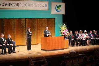 上田 清司知事が会場の壇上で話をしており、知事の横には手話同時通訳者の女性が手話をしており、舞台上には赤い胸章を付けた関係者の方々が並んで椅子に座っている式典の写真