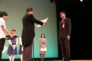 背広を着ている男性が表彰を受けて、市長が賞状を読んでいる写真