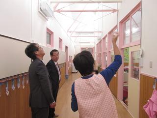 園舎内の廊下の天井に白とピンク色の鉄パイプの柱が補強されているのを保育士の女性が市長と関係者の男性に説明している写真