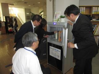 本を殺菌・消毒する機器の扉を開いて、市長と関係者の方々が中の本を見ている写真
