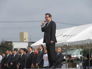 市長が朝礼台の上に立ってマイクを持って話をしており、関係者の方々がテントの前に整列して市長の話を聞いている写真