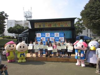 表彰を受けた子ども達が賞状も持って舞台の上に並んでおり、市長とゆるキャラの人形達も一緒に写っている集合写真