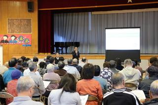 体育館に沢山の住民の方が集まって椅子に座っており、市長が舞台の前でマイクを持って話をしている写真