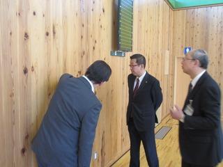 体育館の壁の板を見て、市長が関係者の男性から説明を受けている写真