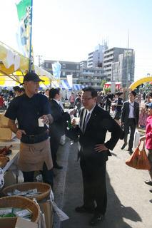 出店の前で黒の帽子に黒のシャツを着た参加者の人と紙コップ片手に会話をしている市長の写真