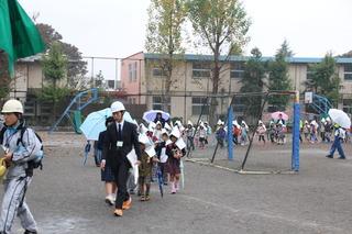 傘をさしたりカッパを着た児童が防災頭巾をかぶり、大人はヘルメットを着用して1列に並び避難している全体を写した写真