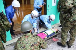 体育館の入口に担架で運ばれた女性が横たわり、その近くに自衛隊職員とヘルメット姿の医師が女性を運ぼうとしている写真