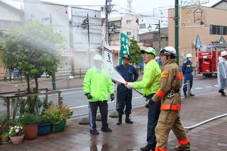 黄色のジャンパーを着た参加者の人が消防職員にホースでの放水指導を受けている写真