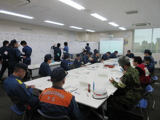 室内の白い大きな机の上にいろんな資料が置かれ消防職員、自衛隊員、参加者などたくさんの人達がいろんな場所で話をしている全体写真