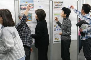 公開討論会で白髪の男性が右手をあげ、その近くに女性や男性が並んでいる写真