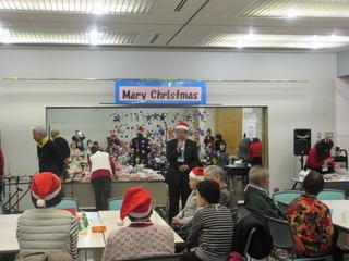 「 Merry Christmas」と横断幕jが貼られクリスマスツリーが飾られている部屋で参加者がサンタの帽子をかぶり挨拶をしている市長をみてる写真