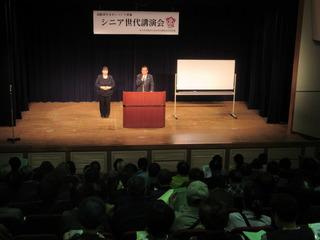 シニア世代講演会で、舞台上の演台に立つ市長と、その横に立つ女性の写真