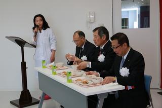 給食を試食する市長と、関係者の横で、女性がマイクを持って話している写真