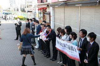 街頭で学生が職員が首に下げている募金箱に募金をしている横で職員が横断幕を掲げて熊本地震義援金募金を呼びかけている写真