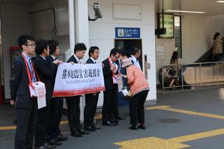 街頭で帽子を被った女性が職員が首に下げている募金箱に募金をしている横で職員が横断幕を掲げて熊本地震義援金募金を呼びかけている写真