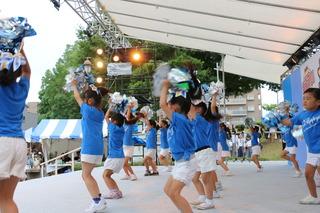 ステージで青い衣装にボンボンを持ってダンスを披露している子供たちをステージ横から写した写真