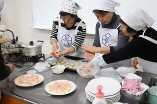 キッチン台で、参加者の女性3名が料理をしており、手で材料を小さく丸めて、鉄板の上に並べている様子の写真