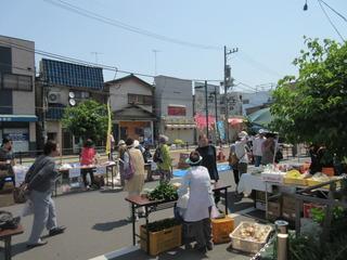 通りにフリーマーケットが開催されて野菜や商品が机に並べられており、通りにはお客さんが行きかっている様子の写真