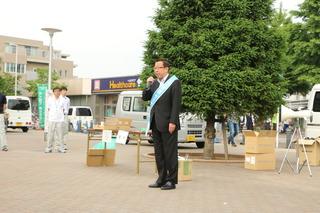市長が水色のタスキを肩にかけて、街頭に立って、マイクを持って話をしている様子の写真