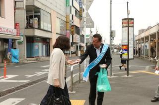 青いタスキを肩にかけている市長が、通りを歩いている女性に、チラシを手渡ししている写真