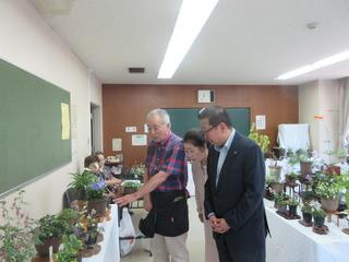 机の上に並べられている鉢植えの植物について、チェックのシャツを着た男性が指を指して説明をしており、その話を市長と女性が興味深く聞いている写真