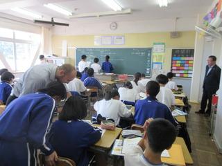 英語の授業が行われており、生徒が3名黒板の前で板書をしており、外国人の先生が後方の生徒の机を覗き込んでおり、市長が前方右側より、生徒の様子を見ている写真