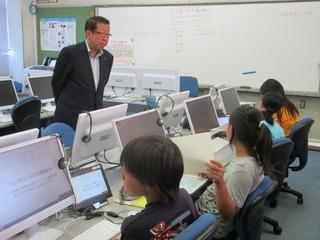 パソコンが並んでいる教室に児童がそれぞれのパソコンの前に座って作業をしており、その様子を市長が子ども達の前に立って見ている写真