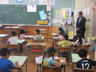 低学年のクラスの授業を参観しており、市長が教室の右前に立って、子ども達が机の上に広げた画用紙に絵を描いている様子を見ている写真