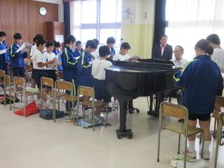 音楽室にてピアノを演奏している先生の周りに生徒が集まって、楽譜を見ながら合唱をしているのを市長が先生の後ろに立って聞いている様子の写真