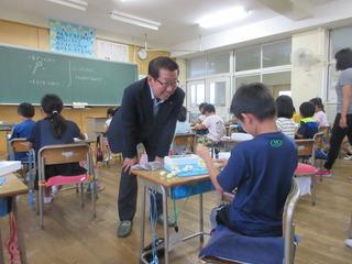 男子児童が机の上で粘土のような物で製作をしており、市長が児童に笑顔で話かけている写真