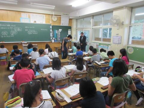 子ども達が教室の席に着いて、机の上にそろばんが置かれて計算をしており、その様子を先生が子ども達の机を回っており、市長が教室の前の入り口に立って様子みている写真
