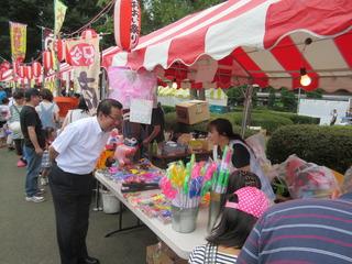 通りに出店が沢山並んでおり、市長がおもちゃが並んでいるお店の女性に笑顔で話をしている写真