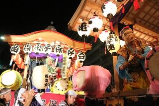 提灯の付いた祭の山車が2機写っており、手前の山車に太鼓が乗っており、お面をかぶった人が太鼓を叩いており、祭りの賑やかな様子の写真
